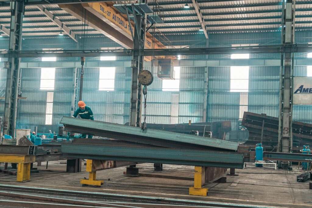 A worker in a helmet manages steel beams in a large factory setting.