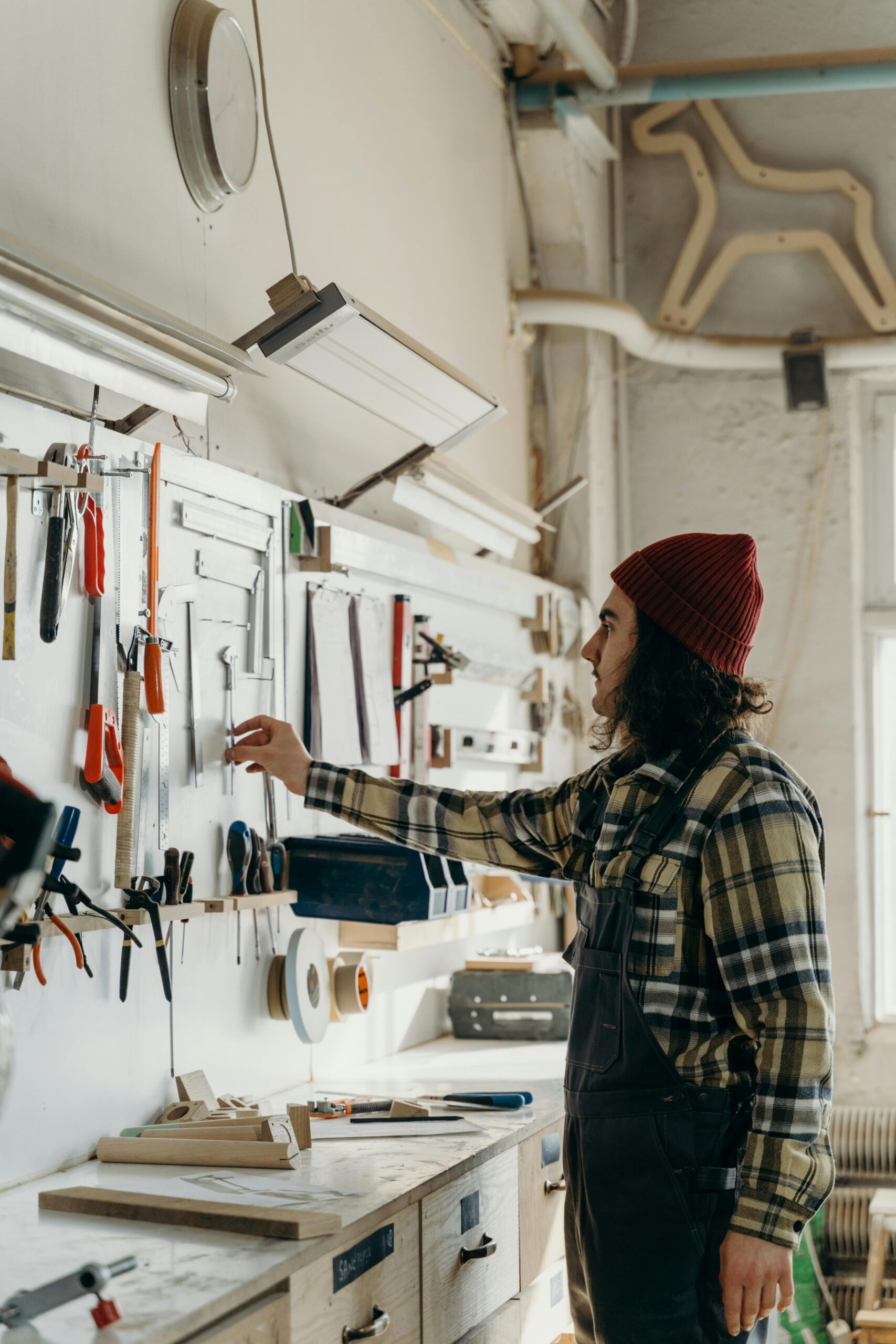 A craftsman in a beanie selecting tools from a workshop wall with organized equipment.